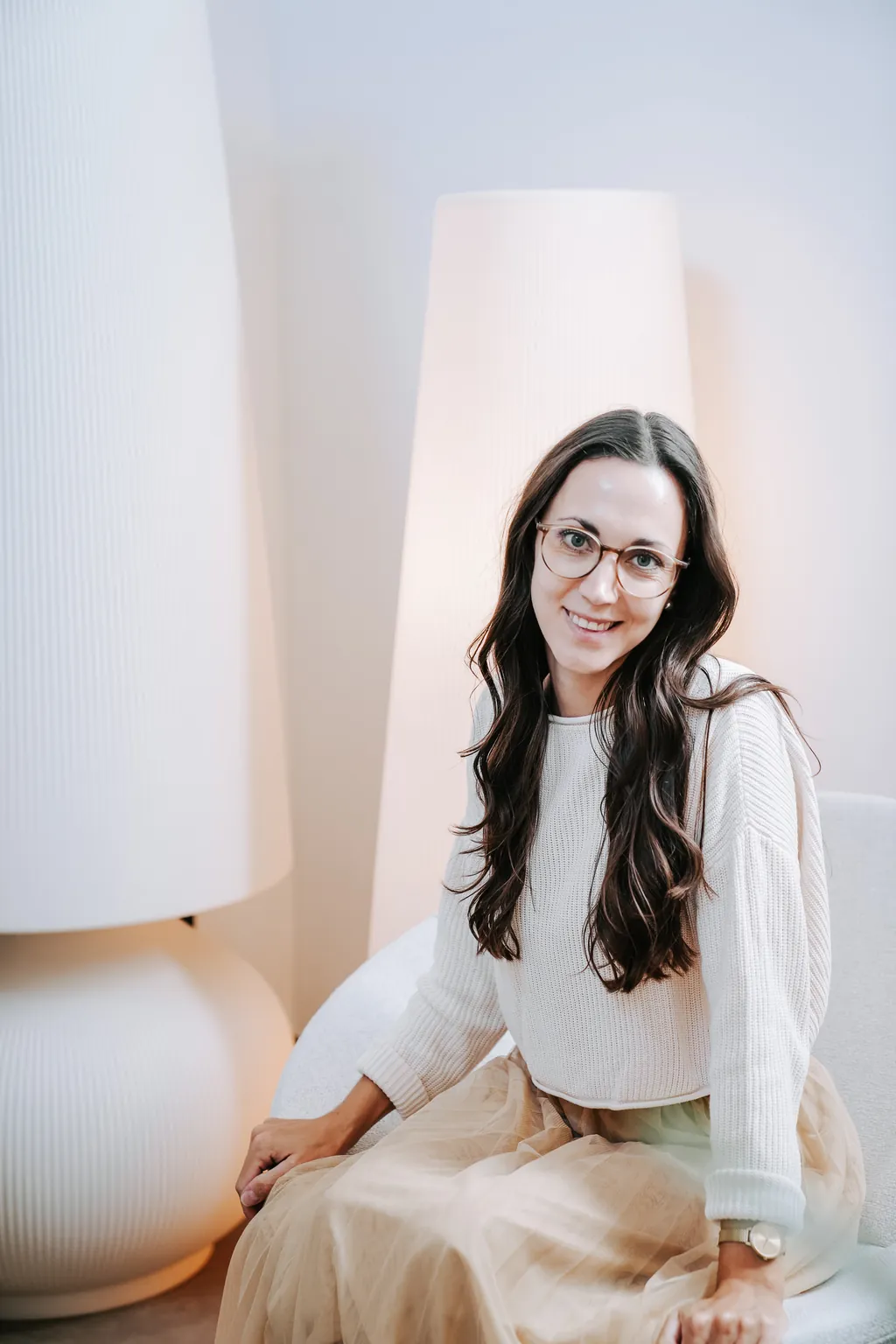 Sophie Täubl mit Brille sitzt lächelnd zwischen zwei hellen Stehleuchten